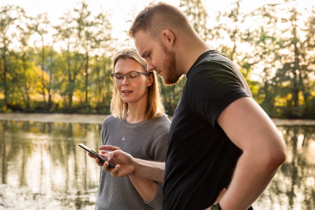 Nainen ja mies seisovat veden äärellä tutkimassa miehen kädessä olevaa matkapuhelinta.