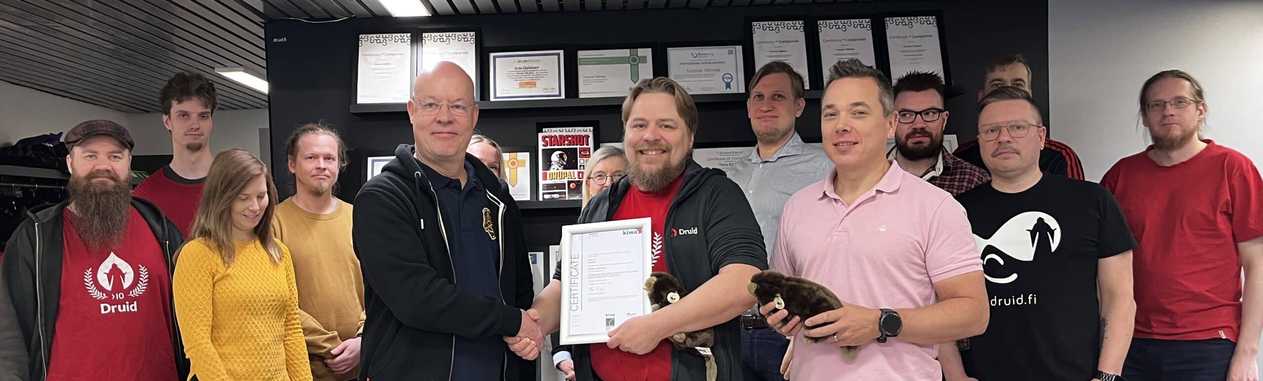 Group of smiling people, some wearing Druid company shirts, standing together indoors. In the center, two men are shaking hands while holding a framed ISO/IEC 27001 certificate, symbolizing an achievement or certification. A variety of certificates can be seen displayed on the wall in the background.