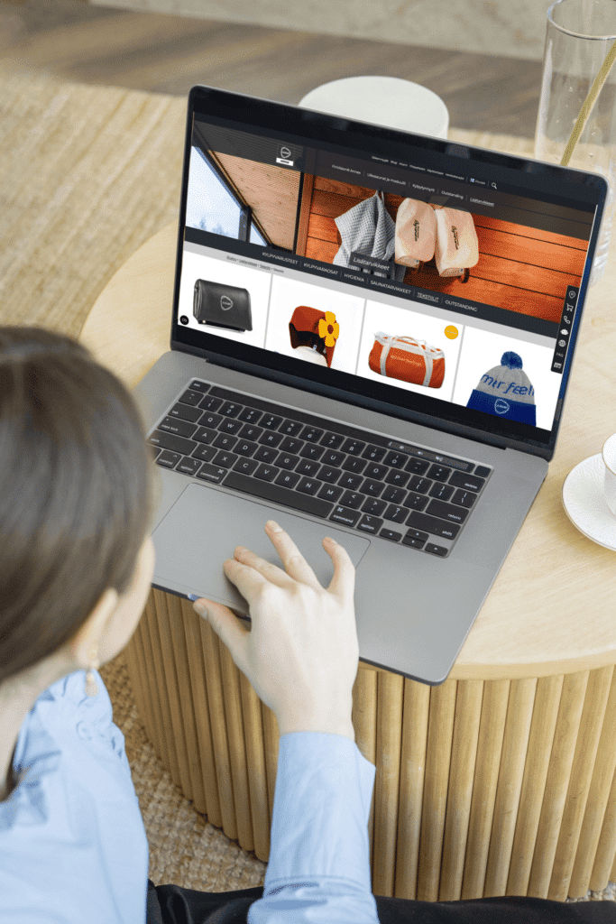 A woman browses an online store on a laptop. The screen displays Kirami's sauna and bathing accessories. The laptop is placed on a round wooden table, with a glass of water and a plate beside it.