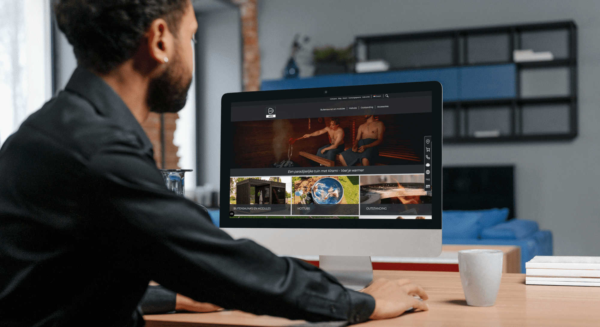 A man browses the Kirami website on a desktop computer in a modern work environment. The screen displays sauna and bathing-related products, featuring an image of men in a sauna and categories such as outdoor saunas, hot tubs, and accessories. On the desk, there is a mug and a glass of water, while a shelving unit and blue-toned interior are visible in the background.