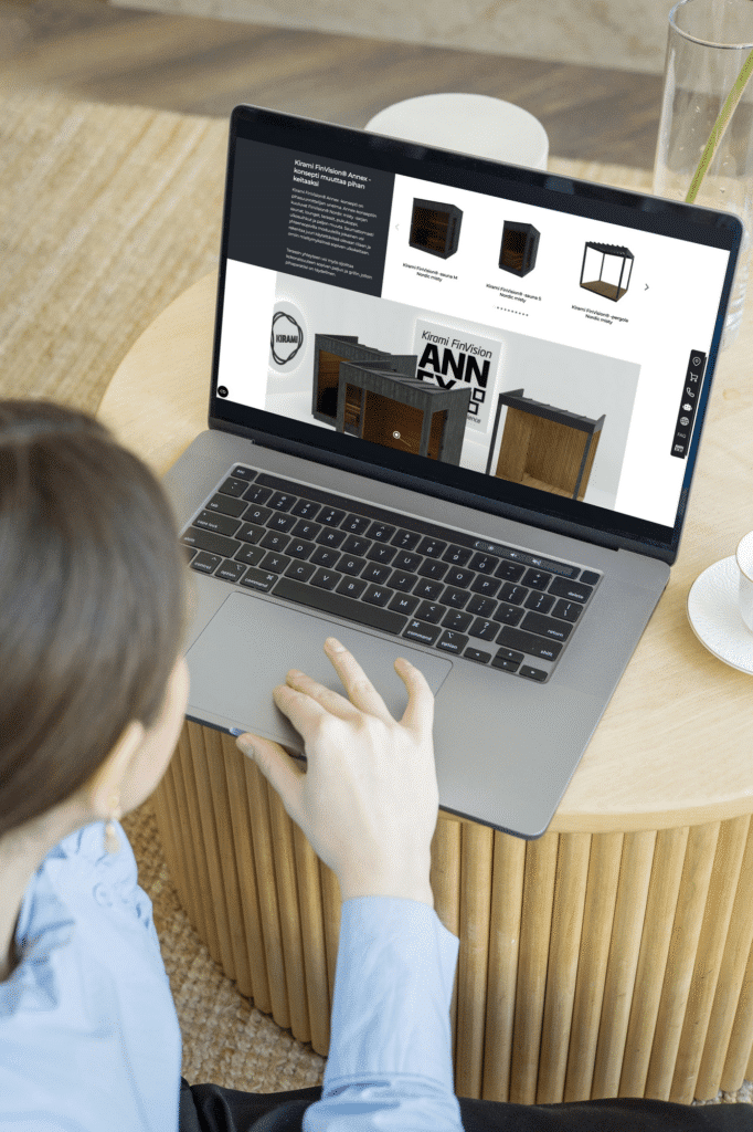 Nainen selaa Kiramin lisävarusteita kannettavalla tietokoneella. Näytöllä näkyy Kiramin Annex-tuotesarjaa. Tietokone on asetettu pyöreälle puupöydälle, ja vieressä on lasi vettä sekä lautanen.