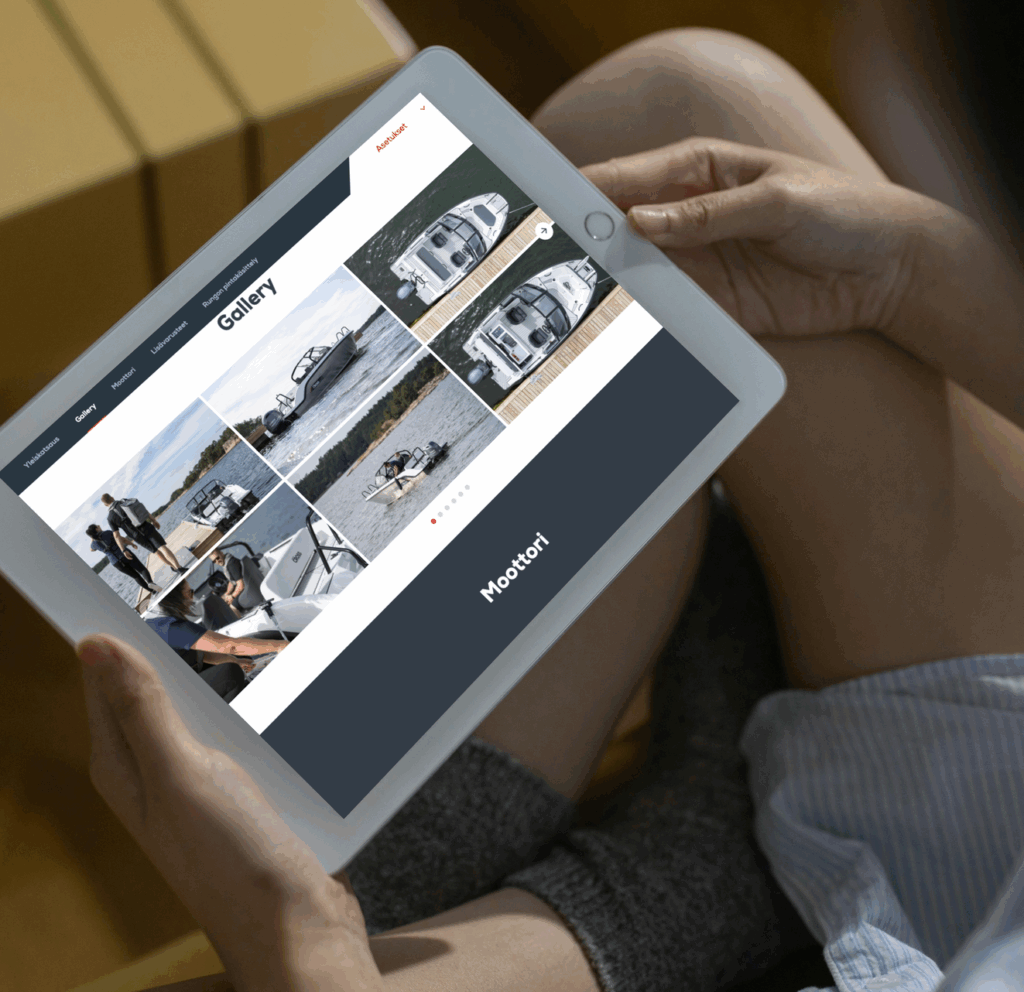 Henkilö selaa tabletilla venegalleriaa, jossa näkyy useita kuvia moottoriveneistä veden äärellä. Näytöllä lukee ‘Gallery’ ja ‘Moottori’. Käyttäjän kädet ja jalat näkyvät kuvassa, taustalla sisätila.